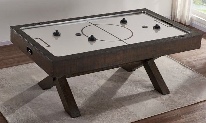 Air hockey table with a dark wooden frame and crossed legs, on a rug near a window.