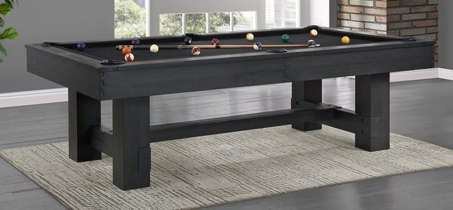 Black pool table on a rug with pool balls set up, in a room with a window.