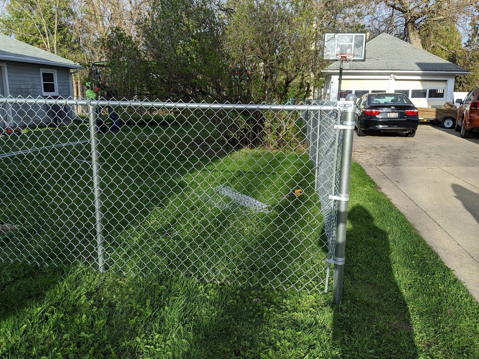 A chain link fence is surrounding a yard with a basketball hoop in the background.