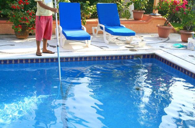 Person cleaning a blue swimming pool beside two blue lounge chairs and potted plants