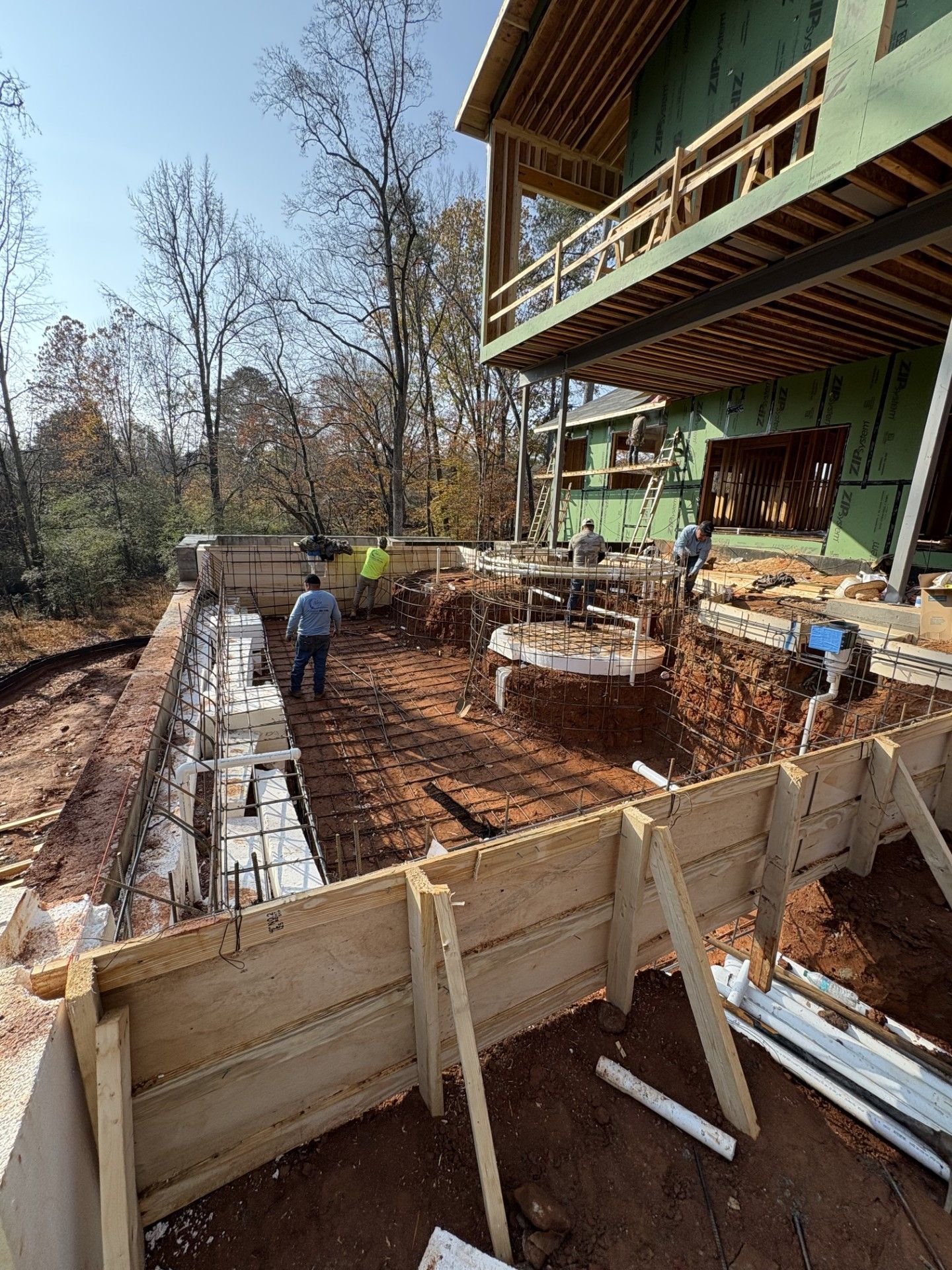Construction workers pouring a concrete patio beside a house with wooden framing and a stone fire pit in a wooded yard