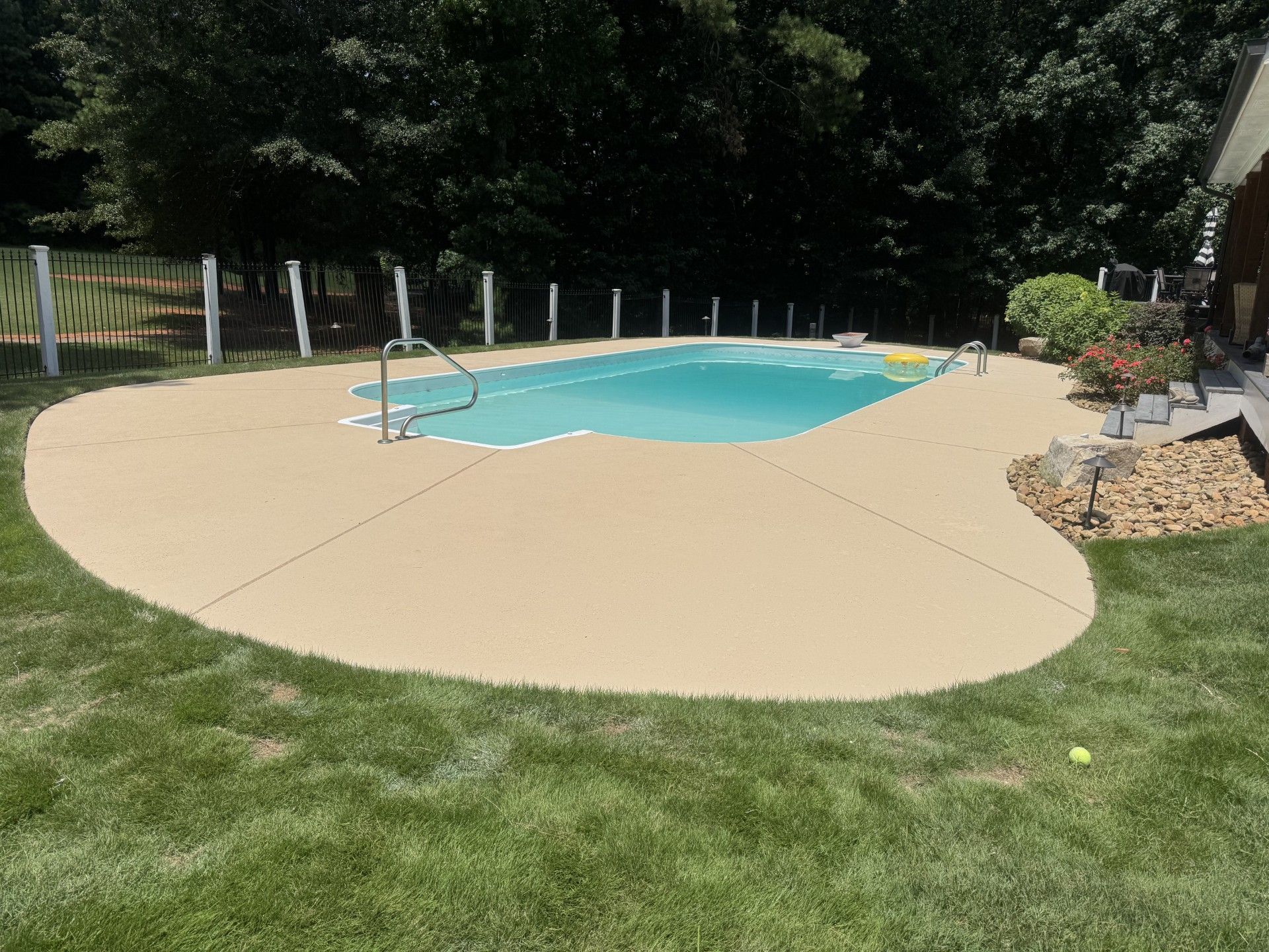 Backyard swimming pool with beige stamped concrete patio and green lawn, surrounded by trees and fence