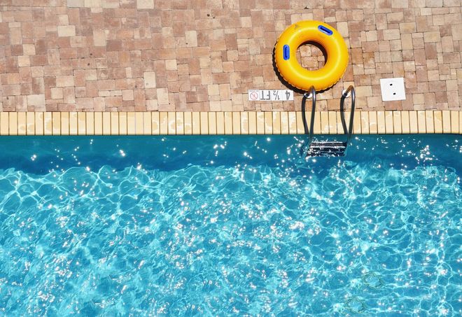 Poolside view with blue water, tan tile deck, and a yellow life ring on the wall.