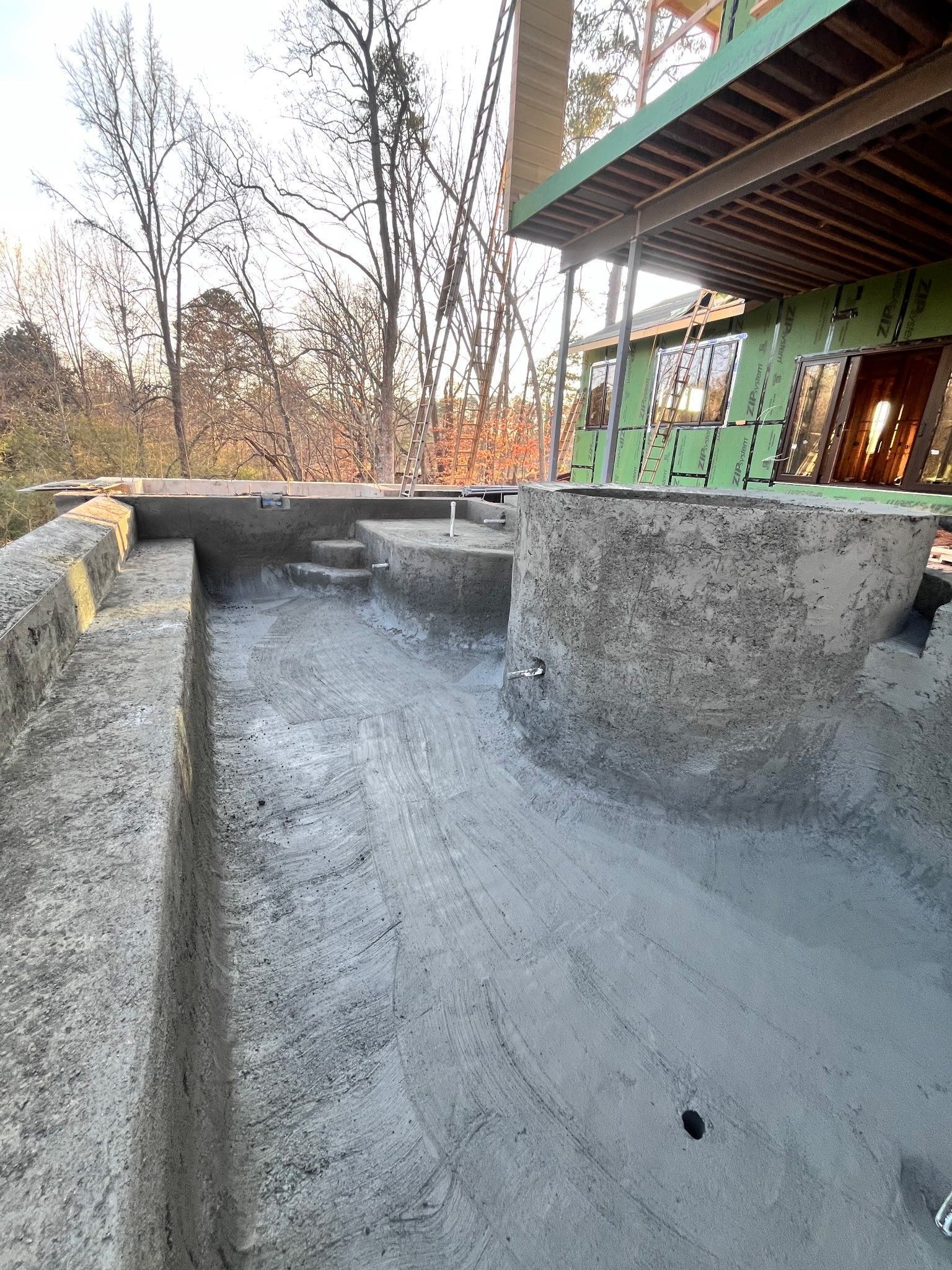 Concrete foundation wall under construction beside a green house, with trees in the background
