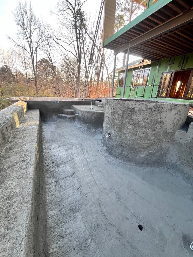 Concrete foundation wall under construction beside a green house, with trees in the background