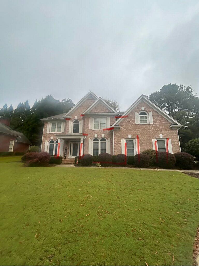 A large brick house with a brown roof is surrounded by trees and bushes.