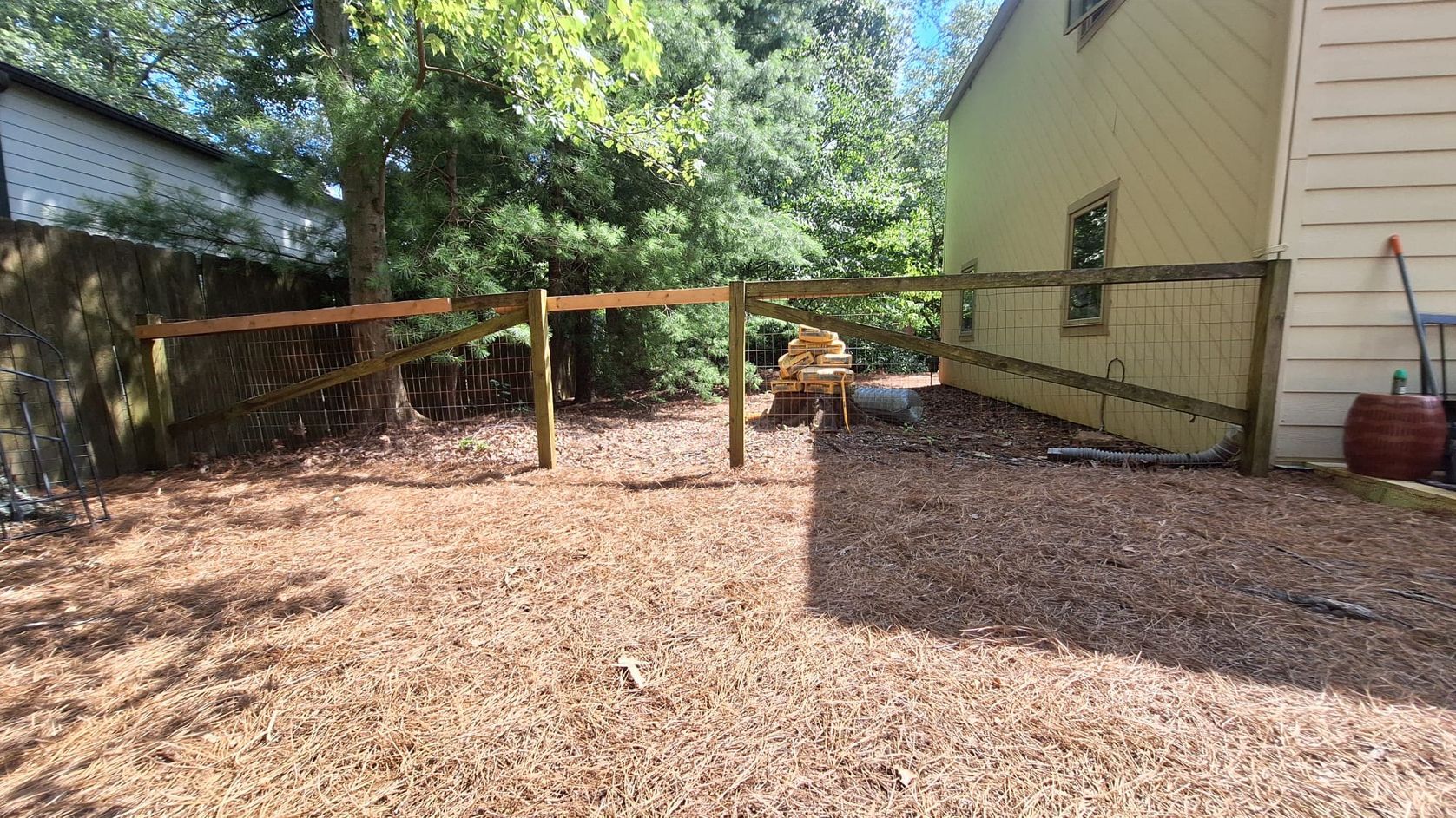 A fenced backyard with wood chips; trees and a light-colored house are in the background.