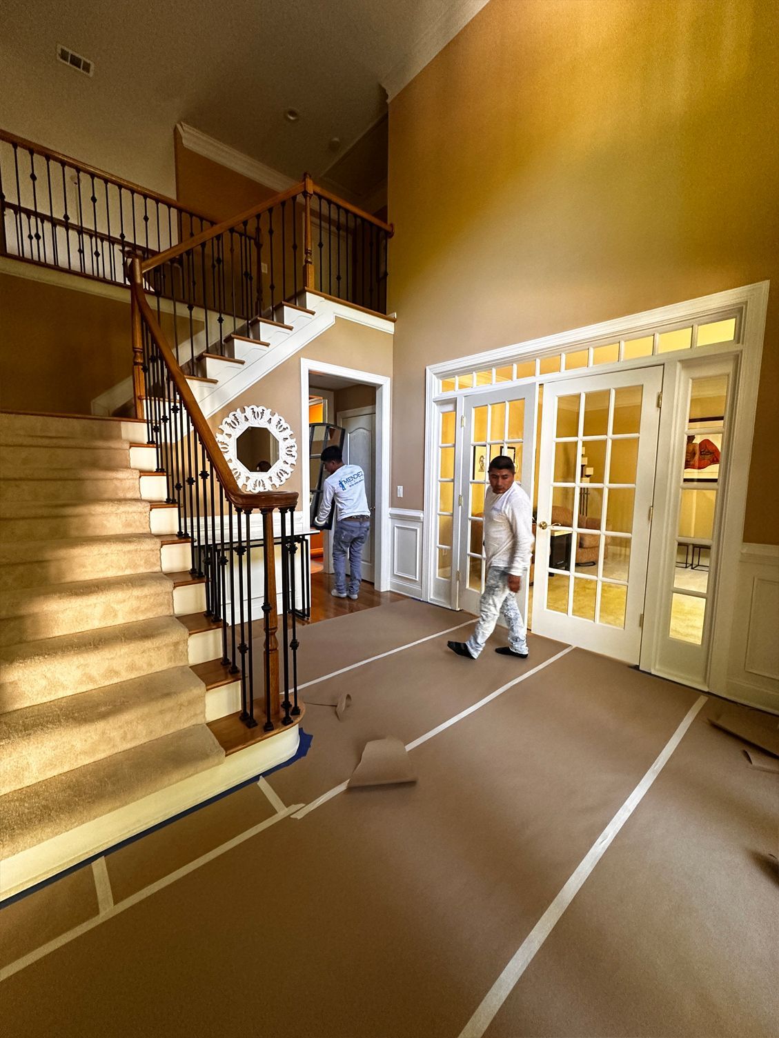 Two painters in a home hallway, one walking, the other near a doorway, both wearing work clothes.