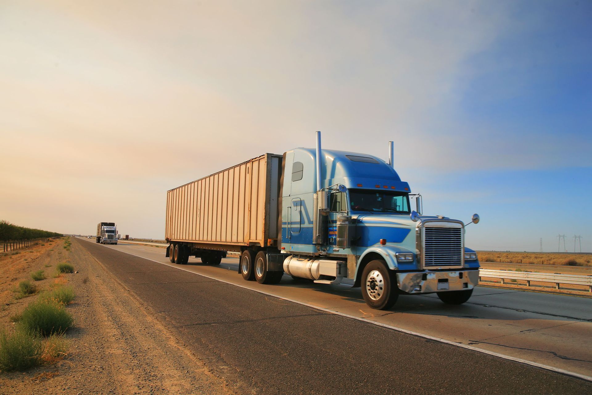 Blue and white semi-truck with a cargo trailer driving on a highway, desert landscape.