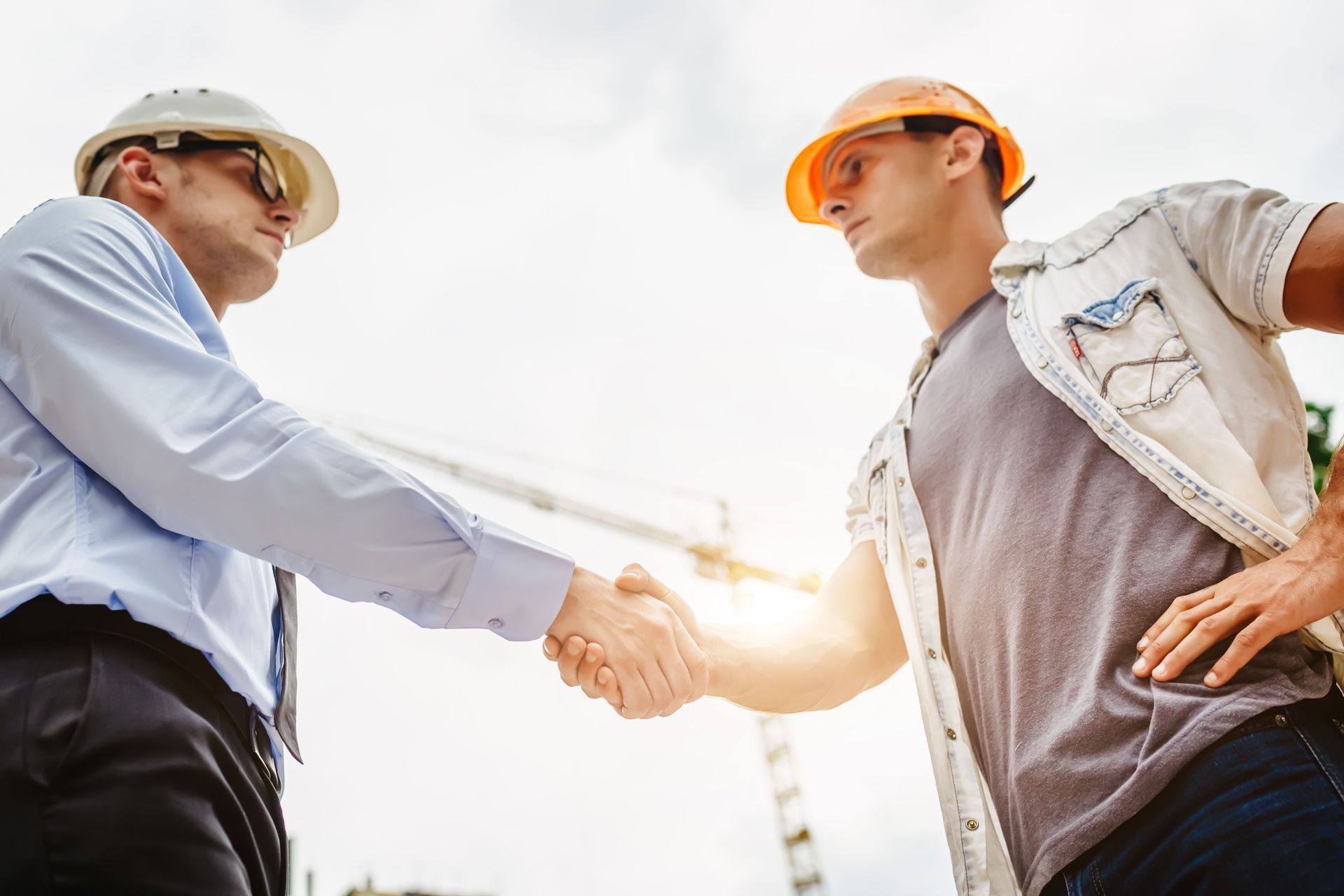 Two people in construction hard hats shaking hands at an outdoor site, with a blurred crane in the background.