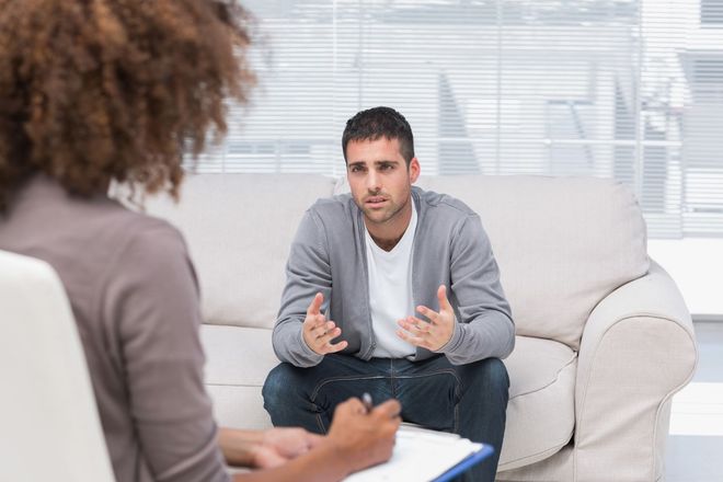 Man speaking during a counseling session on a white couch, with a clipboard in the foreground