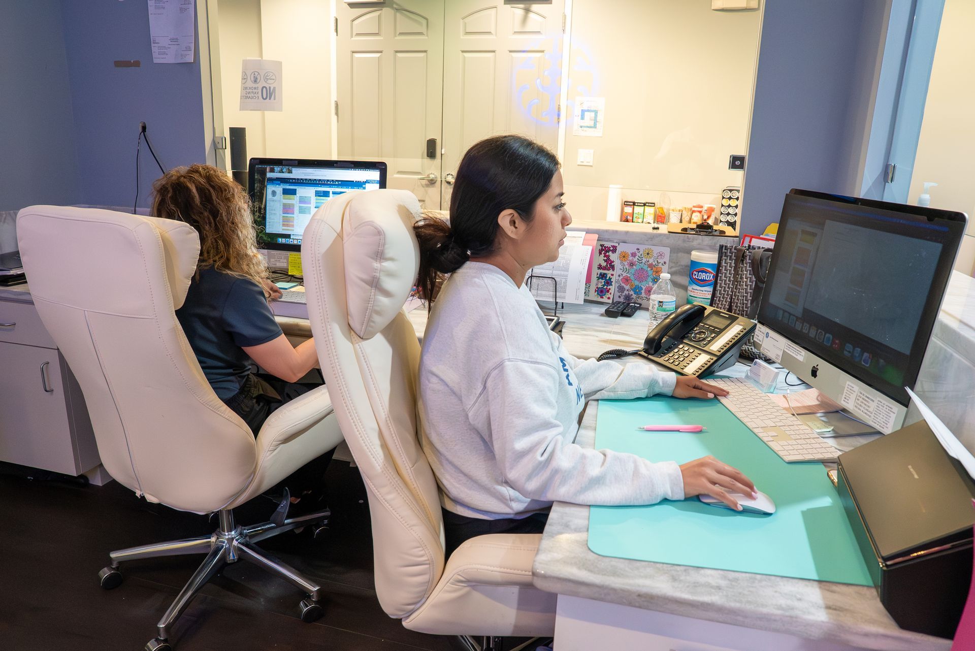 Two people working at computer stations in an office, one typing at a desk with large monitors.