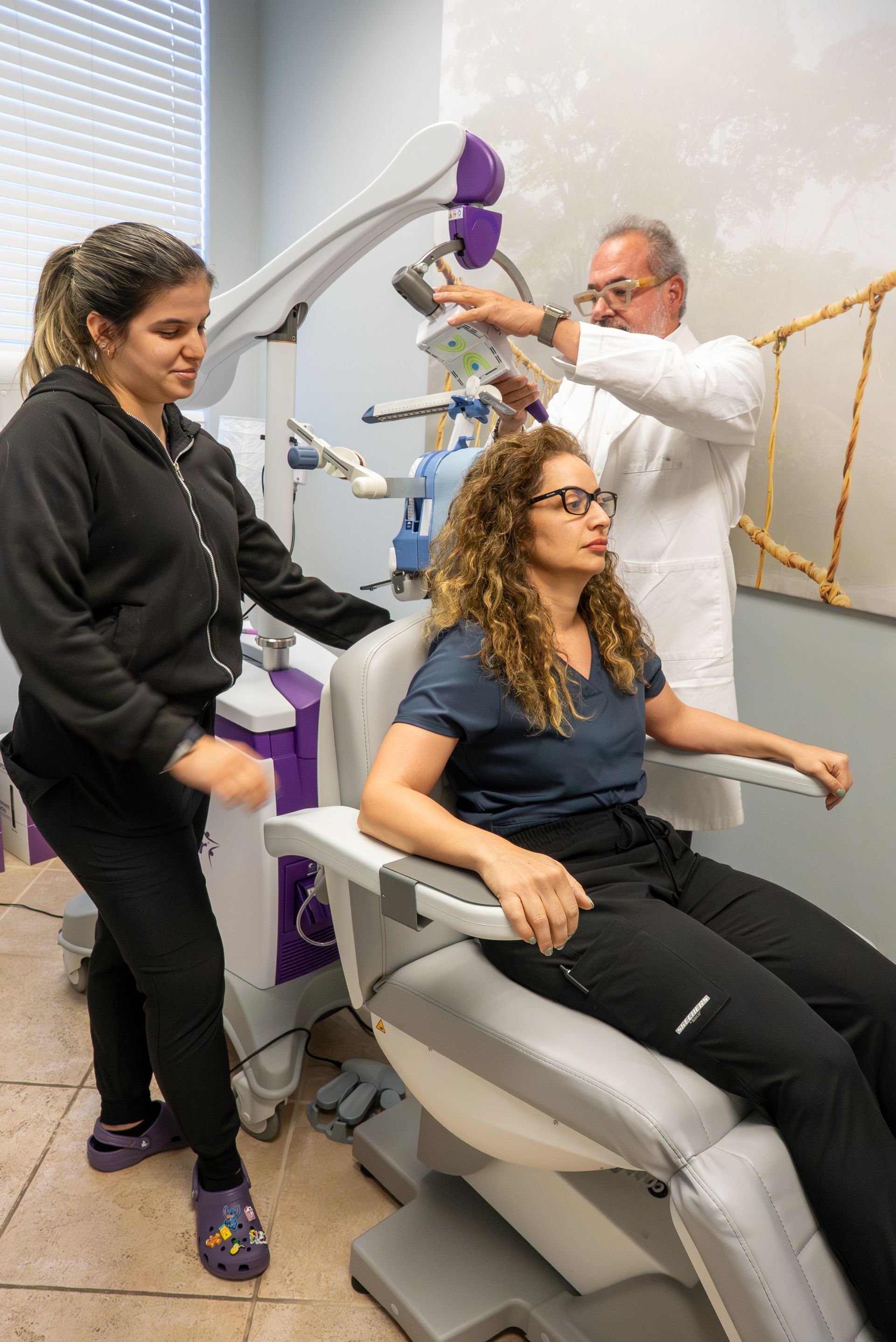 Two clinicians fit a seated woman with a gray robotic exoskeleton in a clinic room