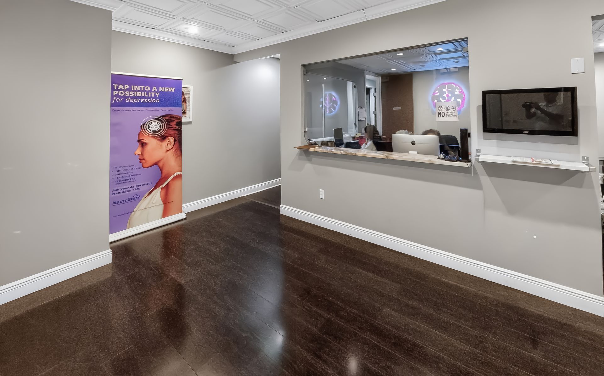 Empty modern office with dark wood floors, gray walls, a reception window, wall-mounted TV, and a purple poster.
