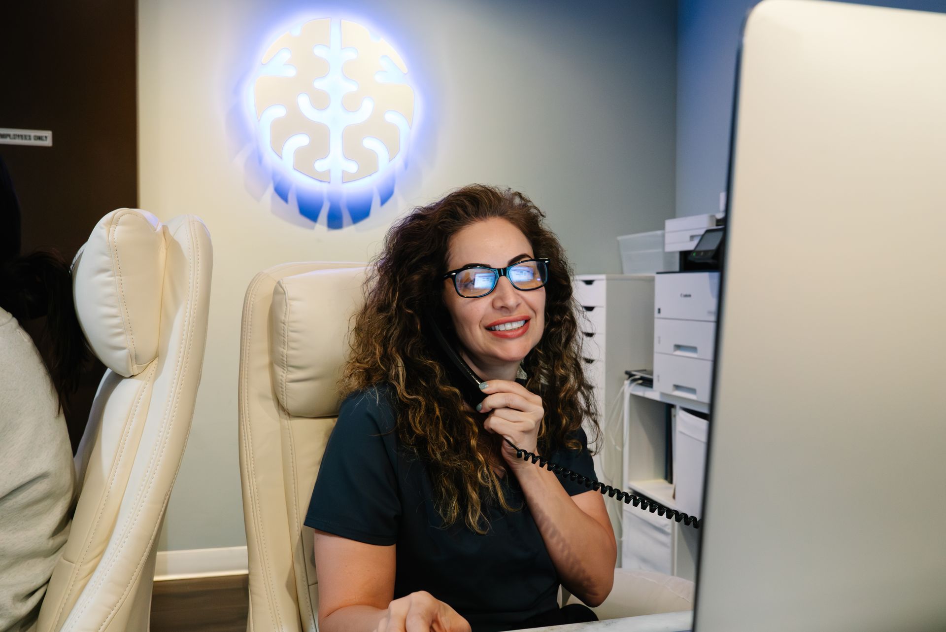 Woman smiling in a clinic chair, wearing glasses, with a glowing brain graphic on the wall behind her