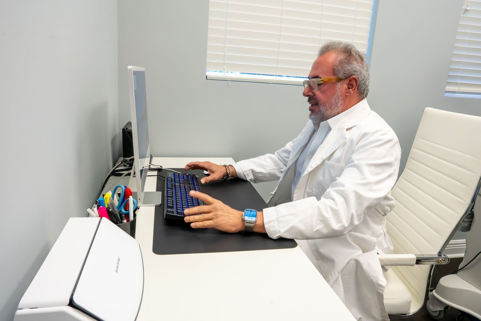 Man typing at a desk in a bright office, wearing a white lab coat and looking at a monitor.