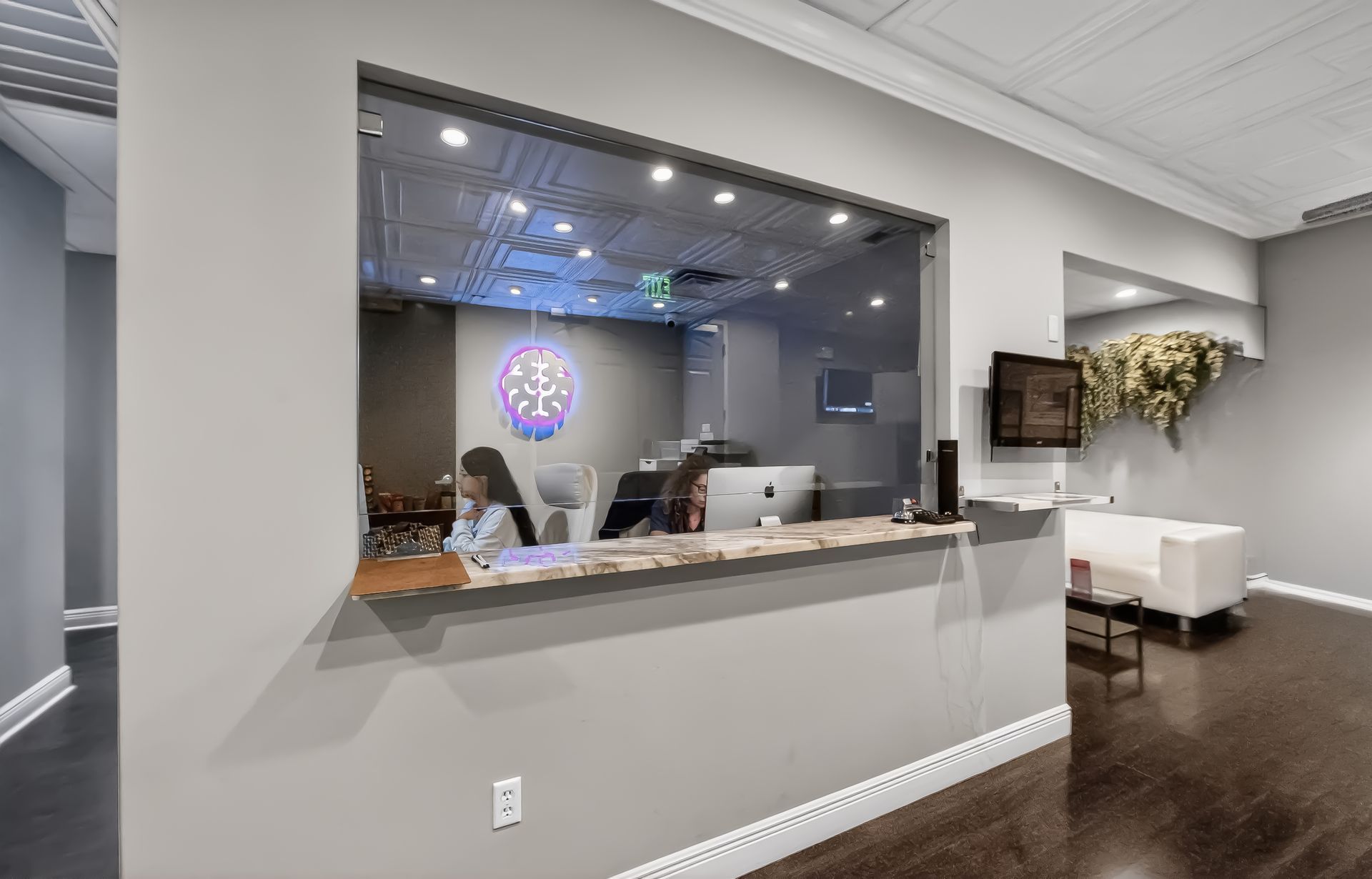 Reception desk and waiting area with white walls, dark wood floors, and a glass window to an office.
