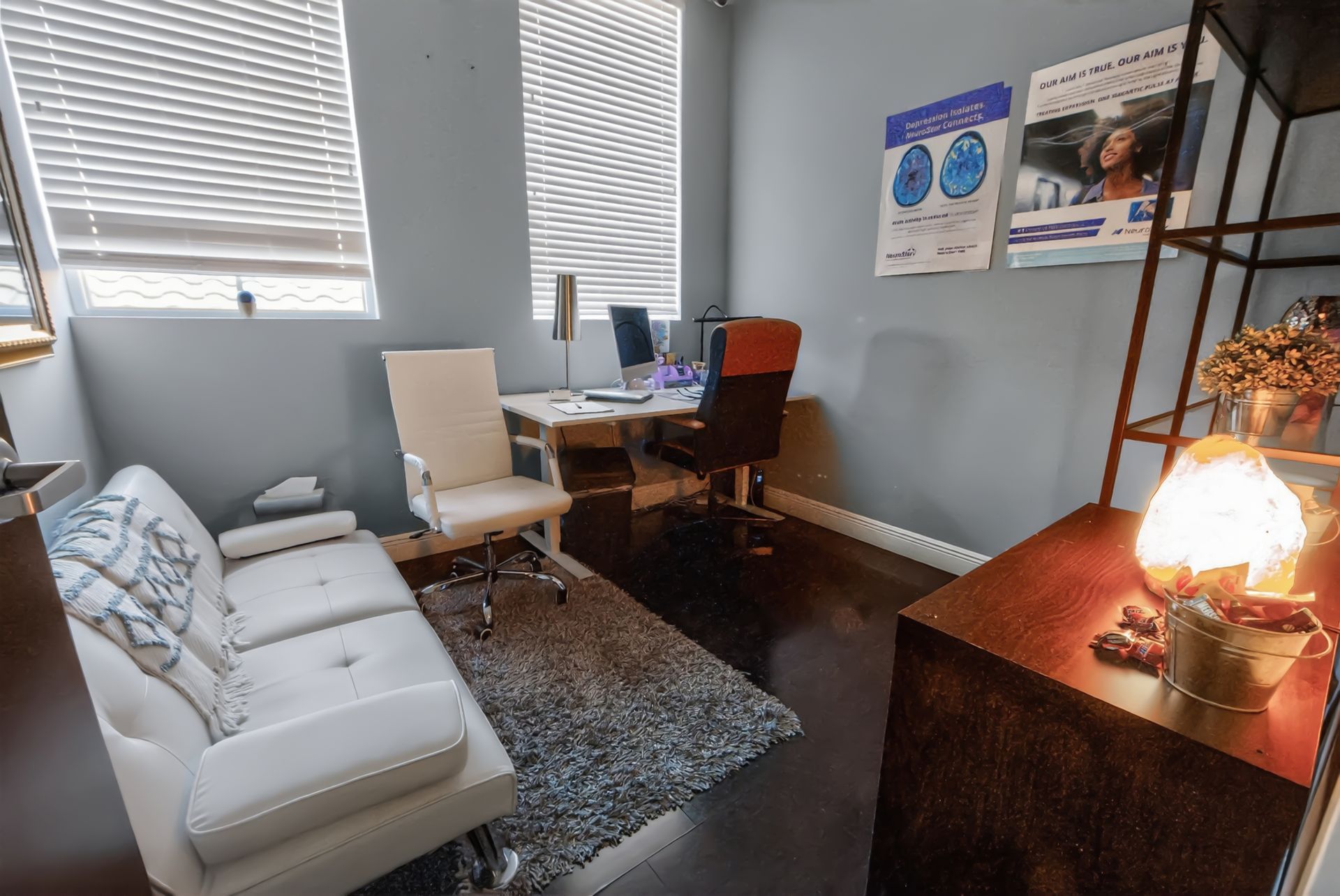 Small office with a sofa, desk, chair, wall posters, and a glowing lamp by the window.