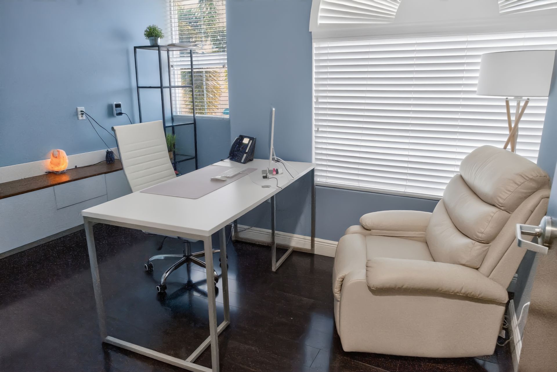 Small office with a desk, chair, beige recliner, lamp, and window blinds in a bright room
