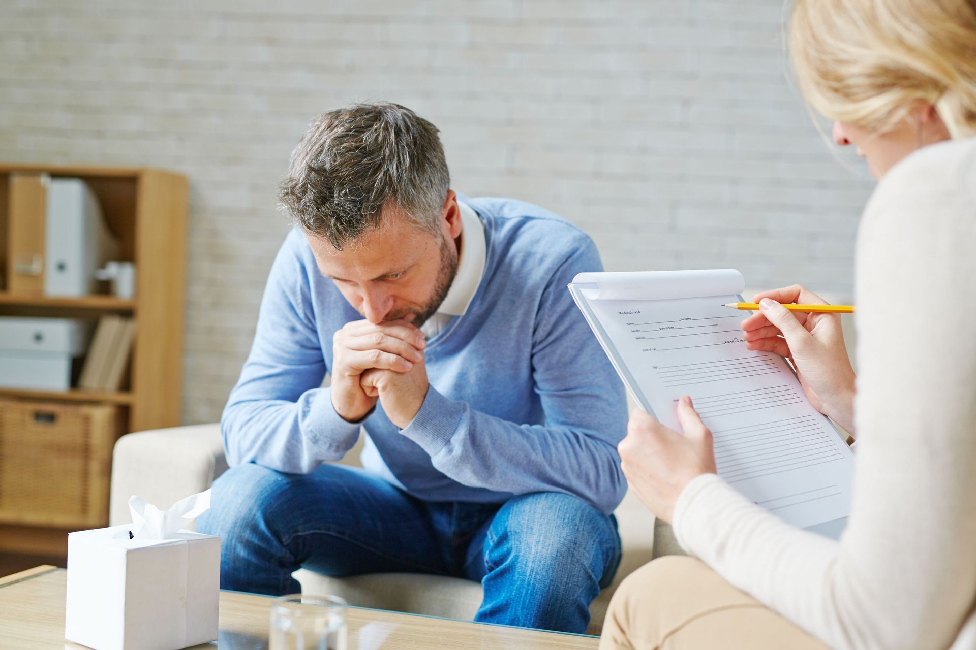 Counselor talking with a distressed man in a home office, holding paperwork and a pencil.