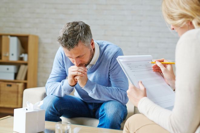 Counselor talking with a distressed man in a home office, holding paperwork and a pencil.