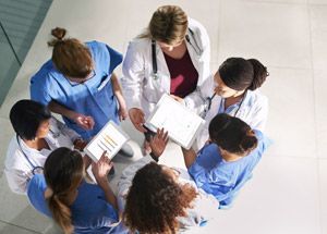 Healthcare team in blue scrubs and white coats reviewing charts in a circle
