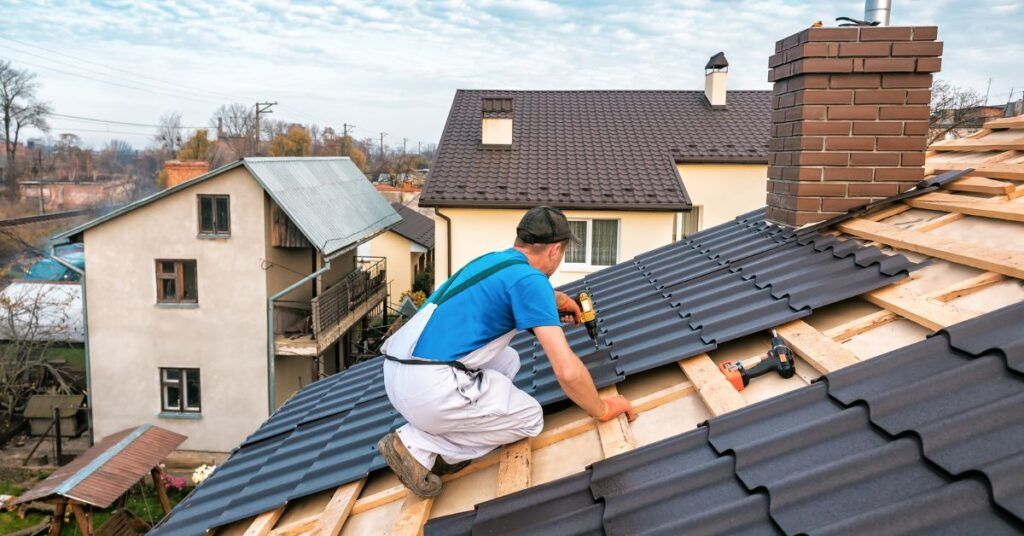 A worker wearing a blue shirt and white overalls uses a power drill to install black roofing tiles on a house.