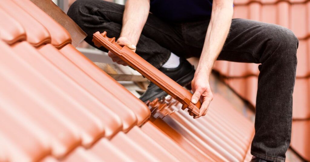 A person wearing dark clothing kneels on a residential roof, carefully installing a single terracotta roof tile.