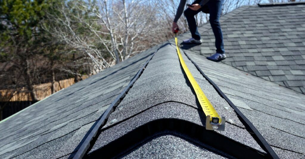A person stands on a roof measuring a ridge line with a yellow tape measure against a bright sky.