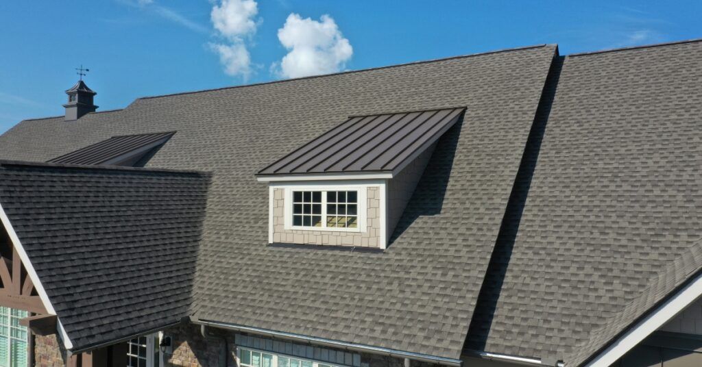 A view of a shingled residential roof with a metal-roofed dormer window under a clear blue sky.