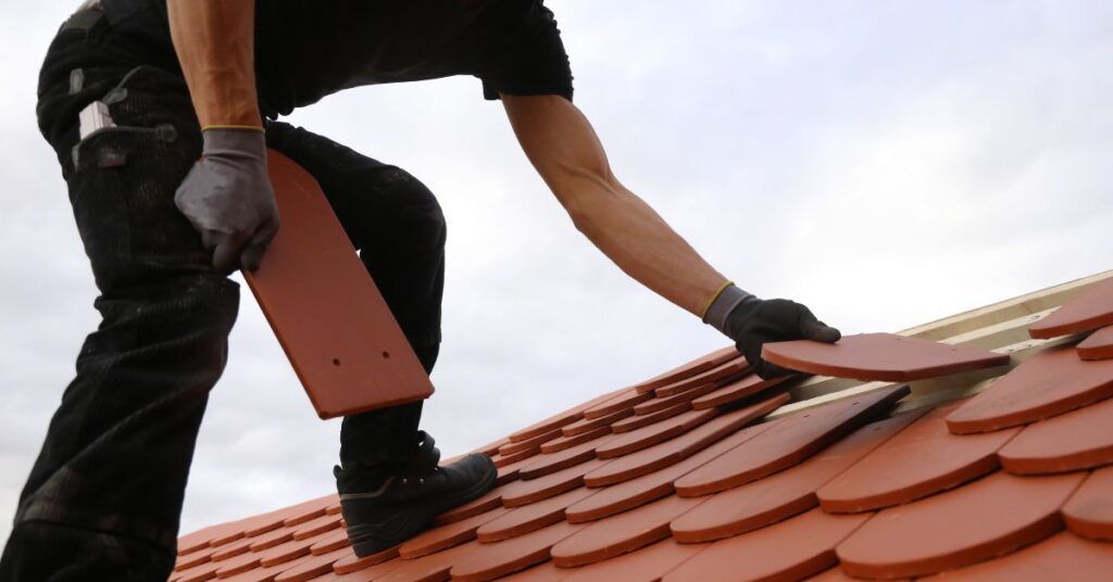 A person wearing gloves places a red clay roof tile onto a roof, surrounded by other installed tiles against a cloudy sky.