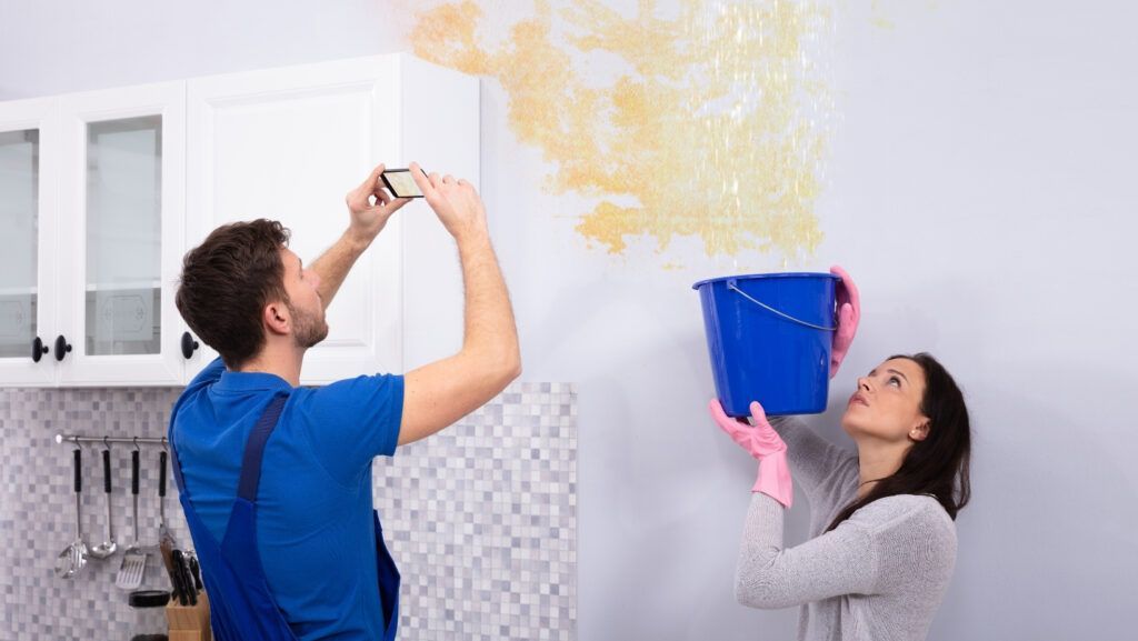 A person in work clothes photographs a water-damaged wall while another person holds a blue bucket under a leak.