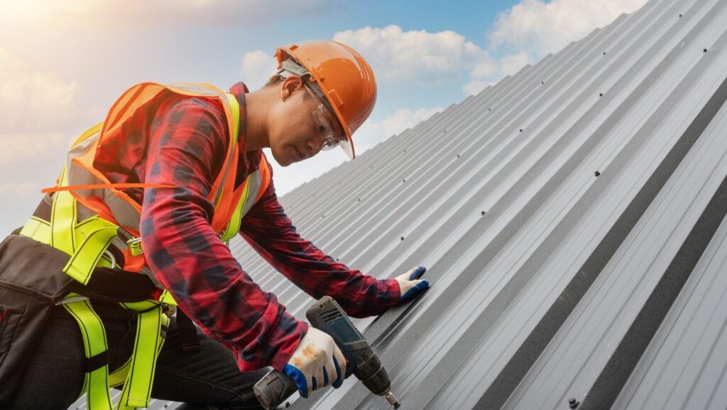 A construction worker in a safety vest and hard hat using a drill to install metal roofing panels on a building.