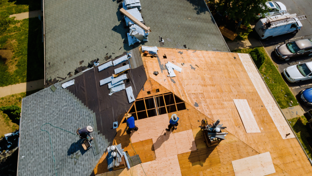 An aerial view of roofers replacing shingles on a house roof, showing exposed wooden deck and new roofing materials.