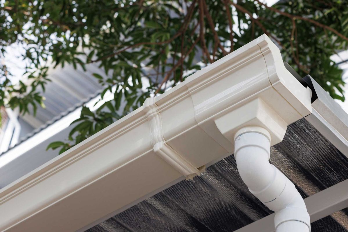 A white gutter system attached to the roofline of a house with a downspout and tree branches in the background.