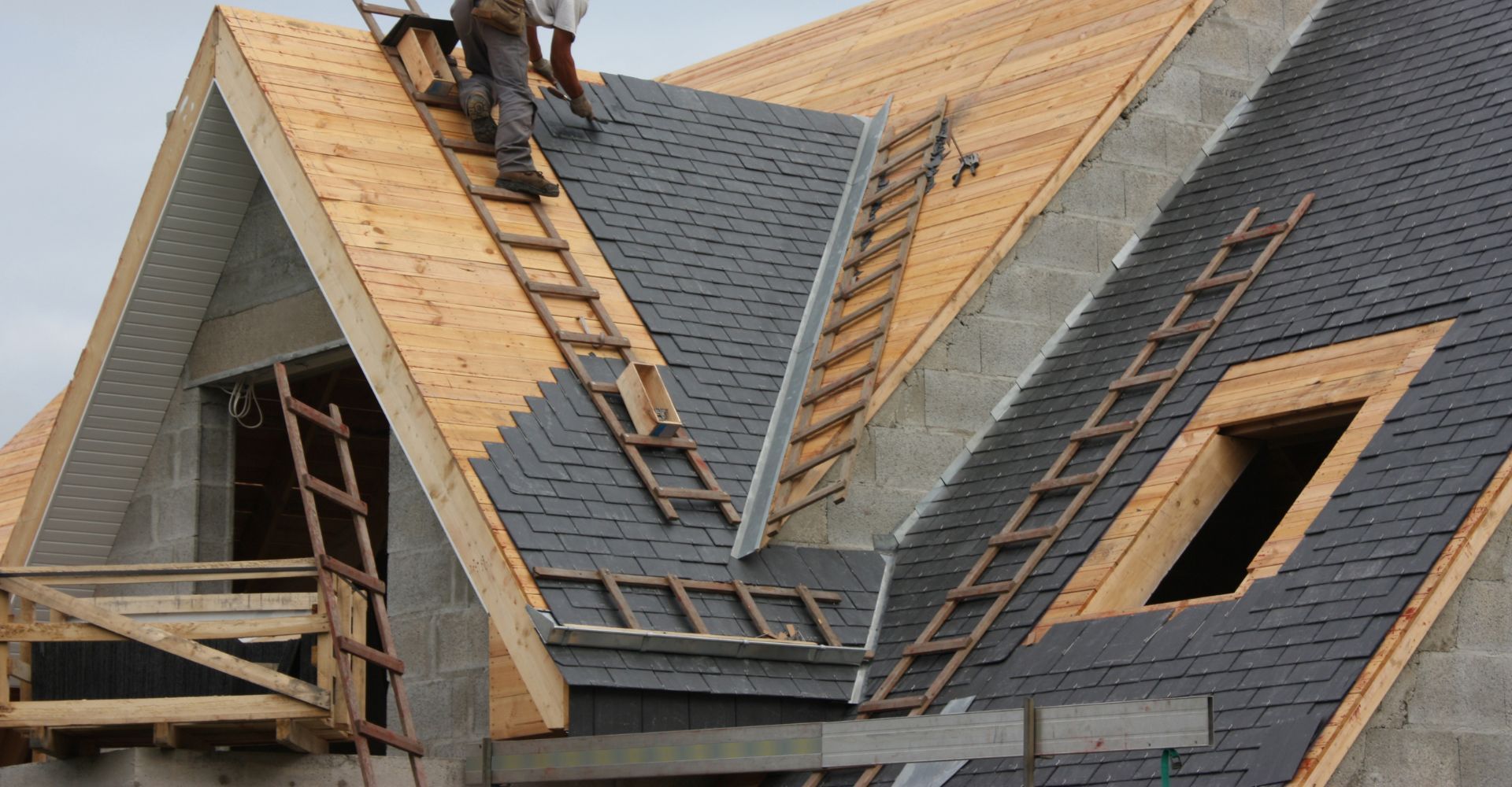 Construction workers on a roof installing dark shingles on an unfinished wood frame house with an attic dormer.
