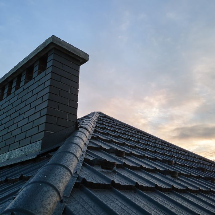 A grey brick chimney stands against a cloudy, sunset sky above a dark, tiled roof.