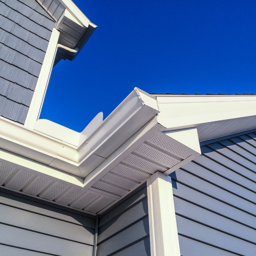 Blue-gray vinyl siding on a home corner with white soffit, fascia, and gutters against a bright blue sky.