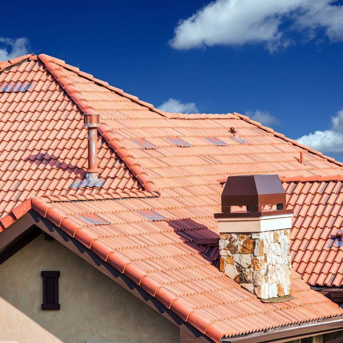 A close-up view of a red terracotta tile roof with a stone chimney and metal vent pipe against a blue sky with clouds.