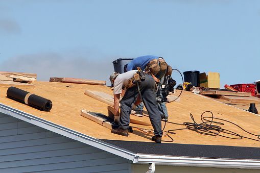 A person wearing a tool belt installs roofing material on a residential house roof under a clear blue sky.