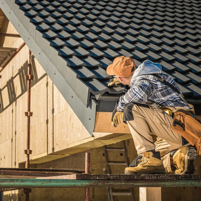 A worker in a plaid shirt and tan work clothes kneels on scaffolding to install black roof tiles on a house frame.