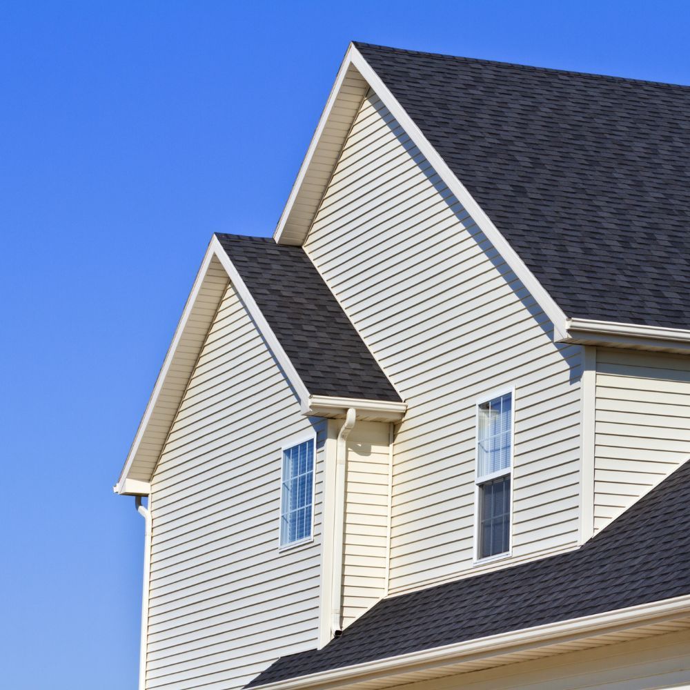 Exterior of a two-story house with cream horizontal siding and a dark shingled roof against a clear blue sky.