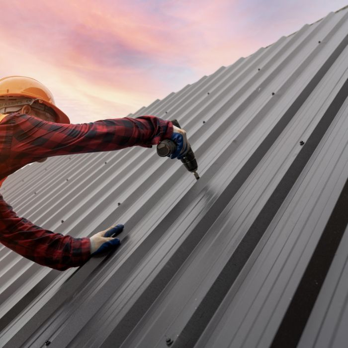 A worker in a plaid shirt and hard hat uses a power drill to install metal roofing panels against a sunset sky.