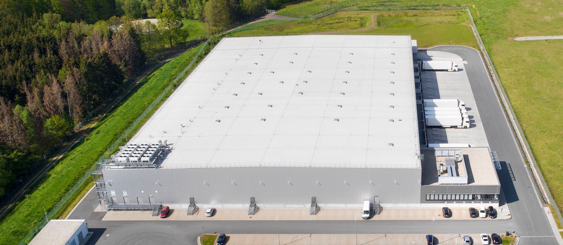 Aerial view of a large rectangular warehouse with a white flat roof, loading docks with trucks, and a parking lot.
