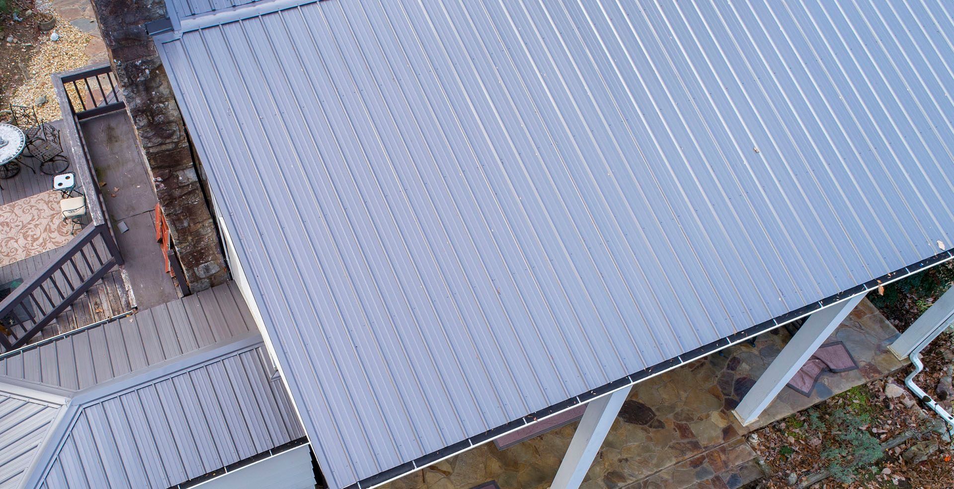 An aerial view of a gray, ribbed metal roof on a house, featuring an adjacent porch with wooden railings and a stone patio.