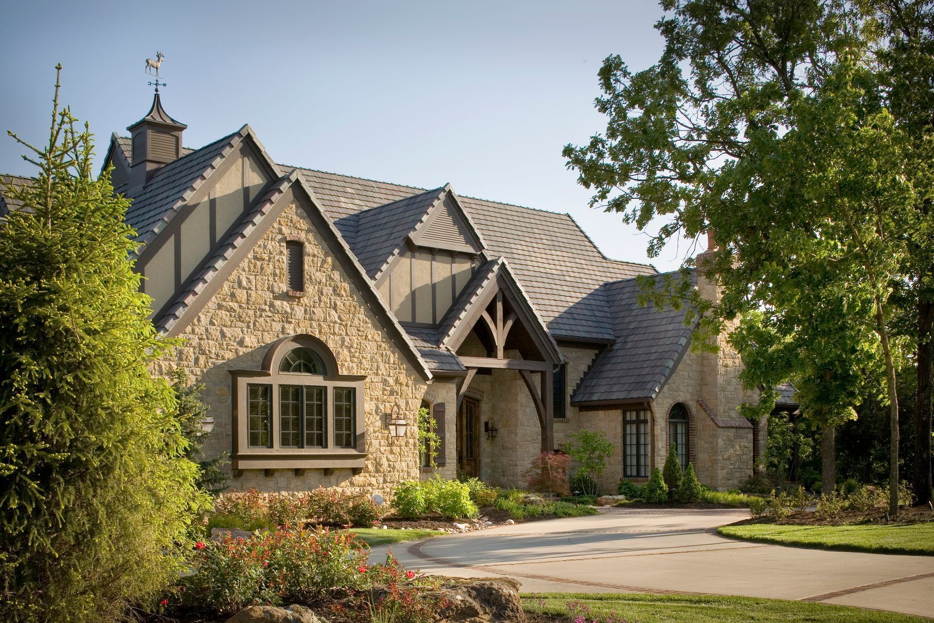 A large, stone-and-timber house with a steep roof, cupola, and arched windows, surrounded by lush landscaping and trees.