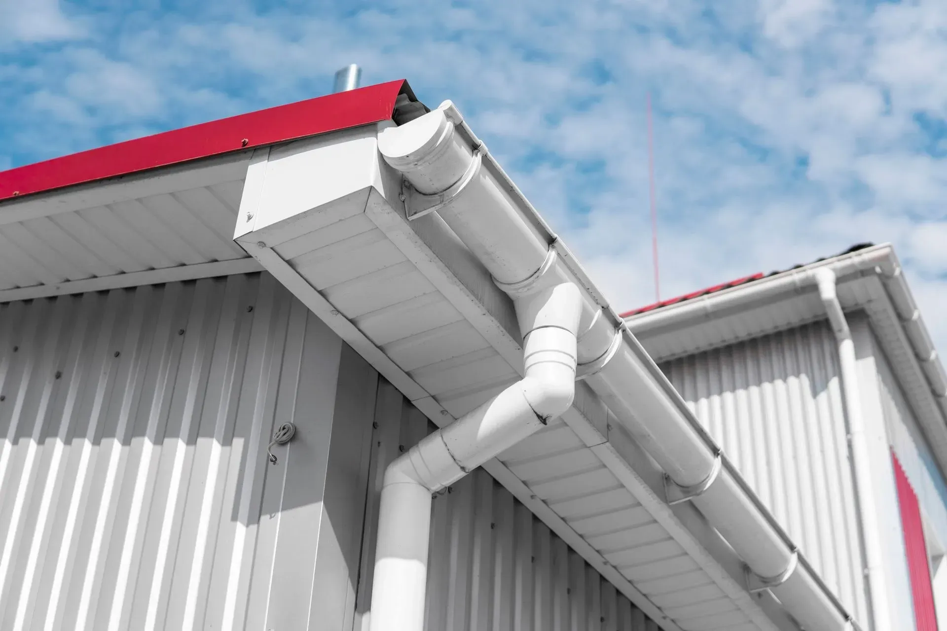 A low-angle view of a white building with a red roof, featuring white gutters and a downspout against a blue sky.