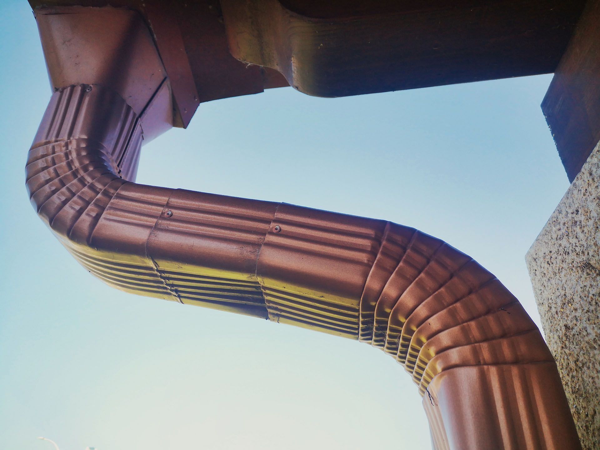 A copper-colored metal downspout makes an S-curve against a blue sky, connecting to a building corner.