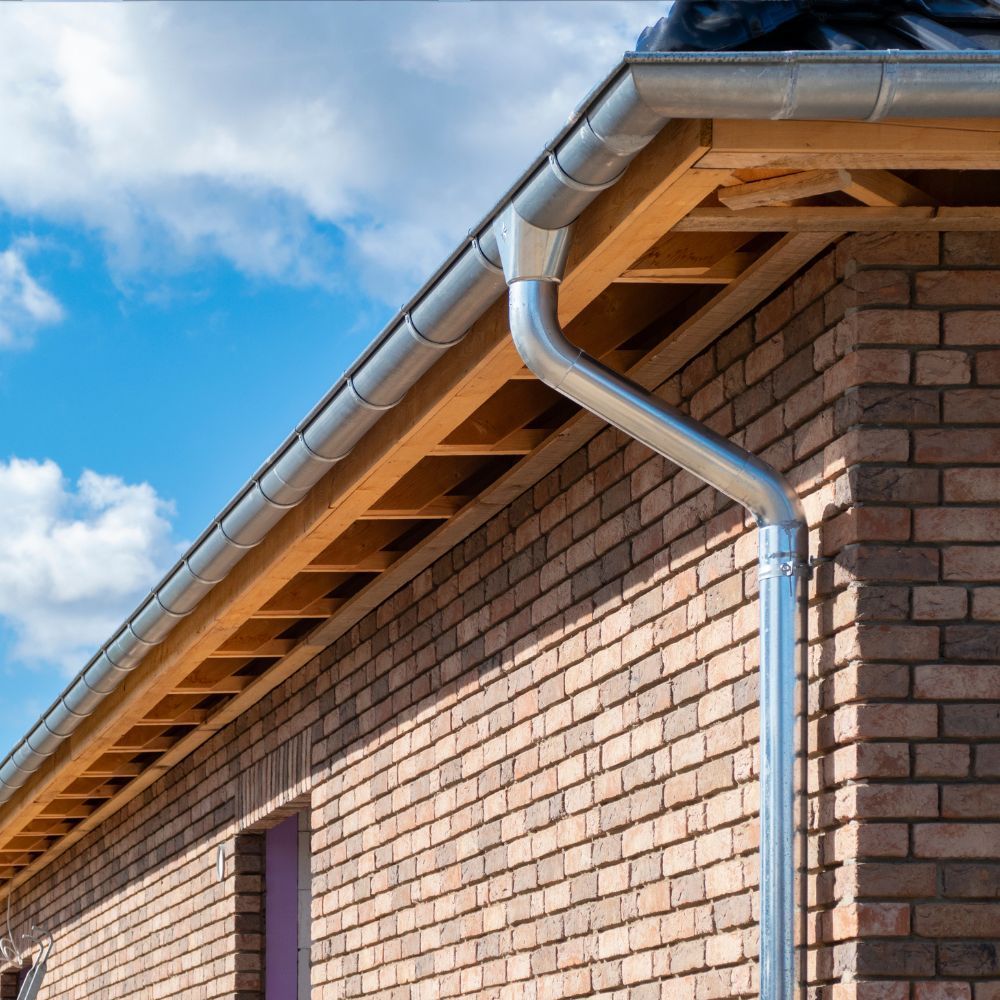 A close-up view of a metal gutter and downspout installed on the brick exterior of a house under a blue, cloudy sky.