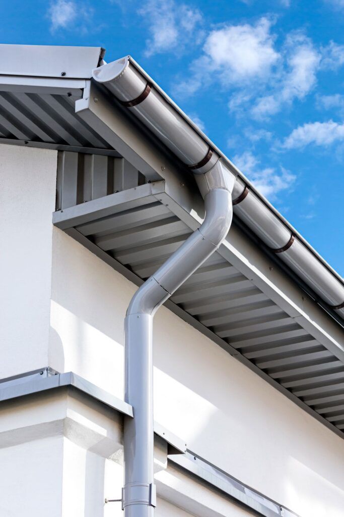 Light gray metal gutter and downspout installed on the corner of a white building against a blue sky.